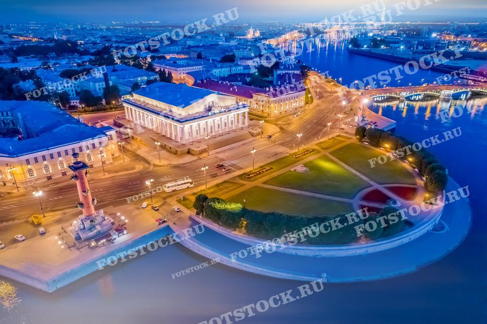 Фотография представляет собой вечерний вид на Стрелку Васильевского острова в Санкт-Петербурге. На переднем плане видно здание Биржи, освещённое в сумерках, и Ростральные колонны, излучающие мягкий свет. Водная гладь Невы отражает ночные огни, и вдалеке видны мосты и контуры города. Фотография идеально подходит для использования в туристических публикациях, в качестве обложки для гидов и книг о Санкт-Петербурге, а также в качестве качественного изображения для печатных и цифровых медиа.
